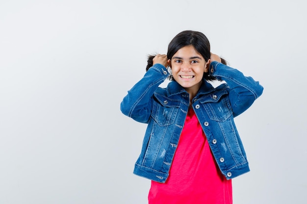 Young girl in red t-shirt and jean jacket tucking hair with hands and looking cute , front view.