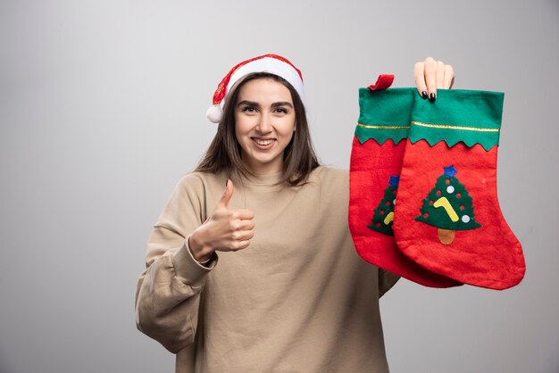 Молодая девушка в шляпе Санты показывает большой палец вверх и держит два рождественских носка.