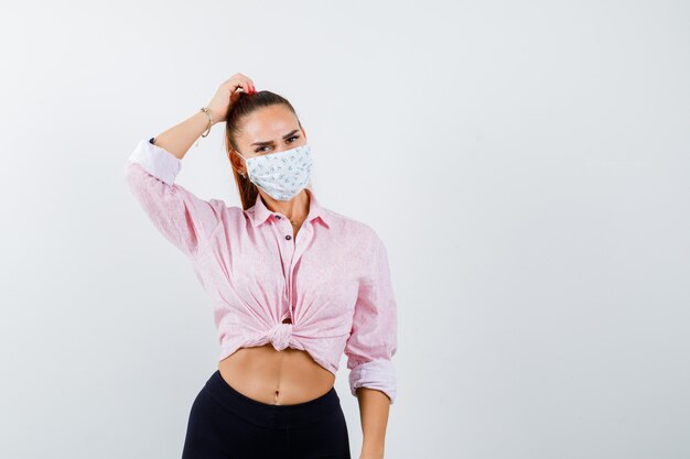 Young girl scratching head in pink blouse, black pants, mask and looking alluring , front view.