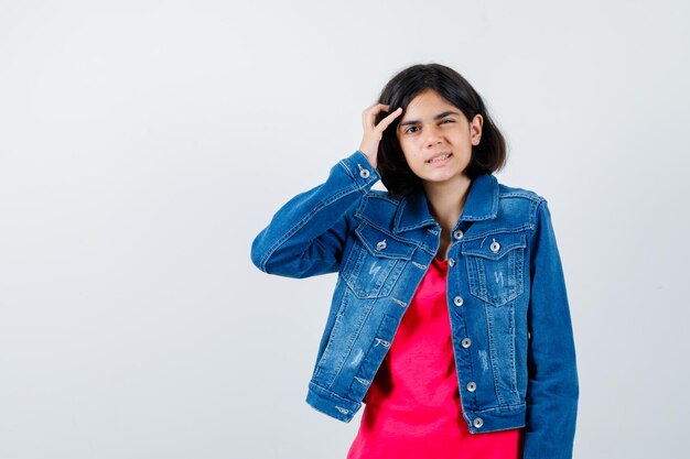 Young girl scratching head, thinking about something  in red t-shirt and jean jacket and looking pensive , front view.