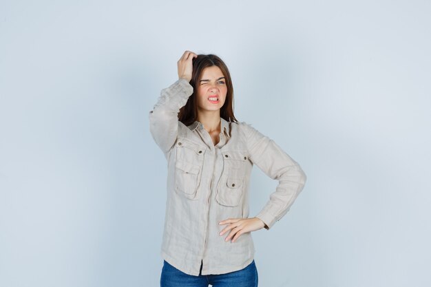 Young girl scratching head while thinking about something, with hand on hip in beige shirt, jeans and looking pensive , front view.