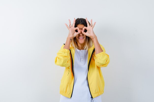 Young girl showing binoculars gesture in white t-shirt , yellow jacket and looking cheery. front view.