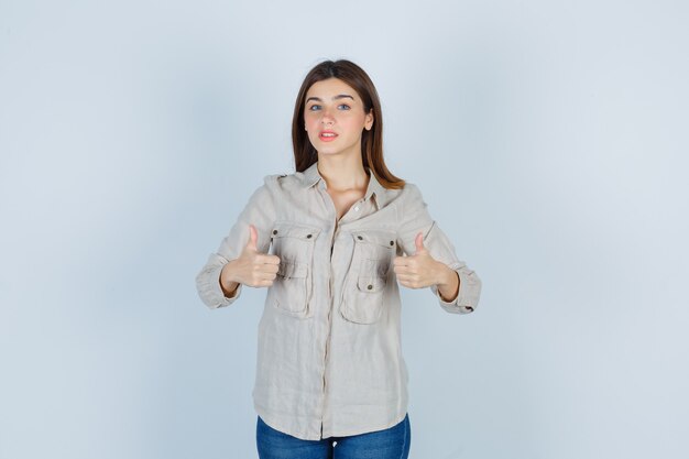 Young girl showing double thumbs up in beige shirt, jeans and looking serious , front view.