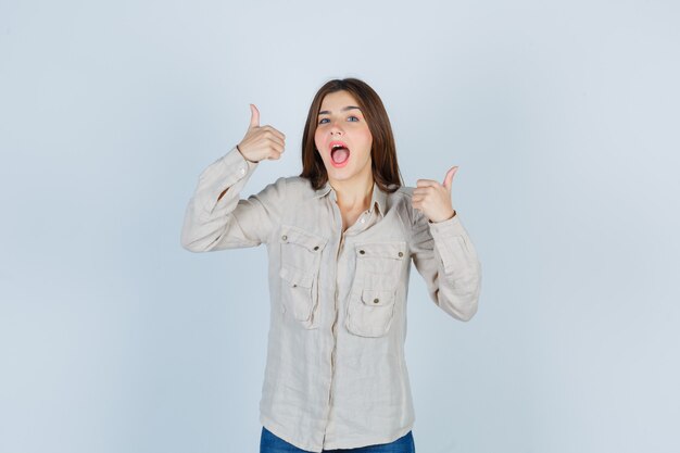 Young girl showing double thumbs up, sticking tongue out in beige shirt, jeans and looking amused. front view.