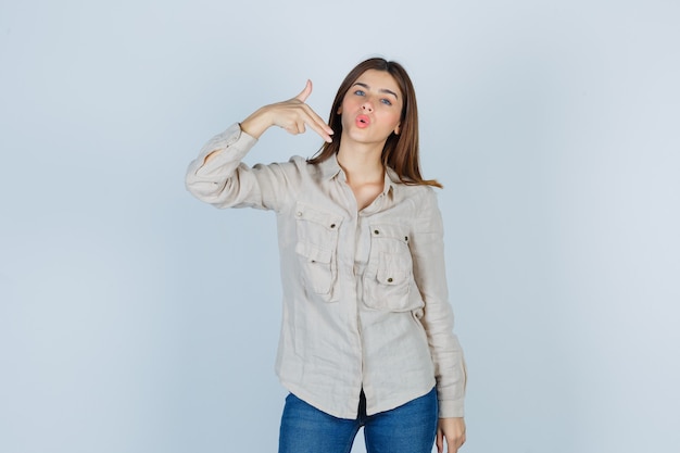 Young girl showing gun gesture in beige shirt, jeans and looking cute. front view.