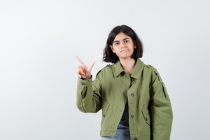 Young girl showing gun gesture in grey sweater, khaki jacket, jean pant and looking optimistic , front view.