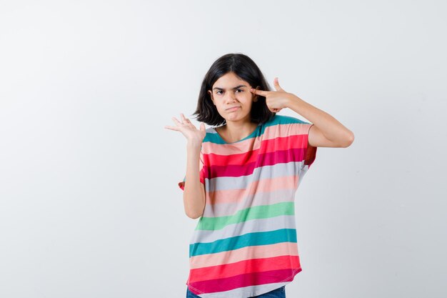 Young girl showing gun gesture near head, stretching left hand in colorful striped t-shirt and looking displeased , front view.