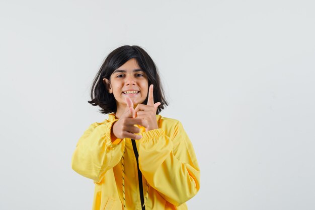 Молодая девушка показывает жесты пистолетом обеими руками в желтой куртке бомбардировщика и выглядит счастливой.
