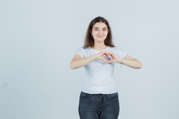 Young girl showing heart gesture in t-shirt, jeans and looking happy , front view.