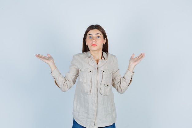 Young girl showing helpless gesture in beige shirt, jeans and looking puzzled. front view.