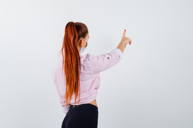 Young girl showing hold on a minute gesture, turning back in pink blouse, black pants, mask and looking alluring , front view.