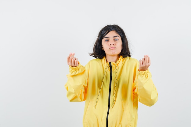 Молодая девушка показывает жест денег в желтой куртке-бомбардировщике и выглядит серьезно