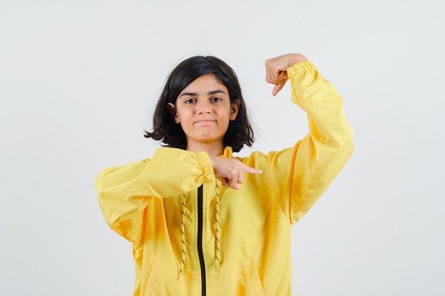 Young girl showing muscles and pointing to it with index finger in yellow bomber jacket and looking happy.
