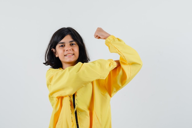 Молодая девушка показывает мышцы в желтой куртке бомбардировщика и выглядит счастливой.