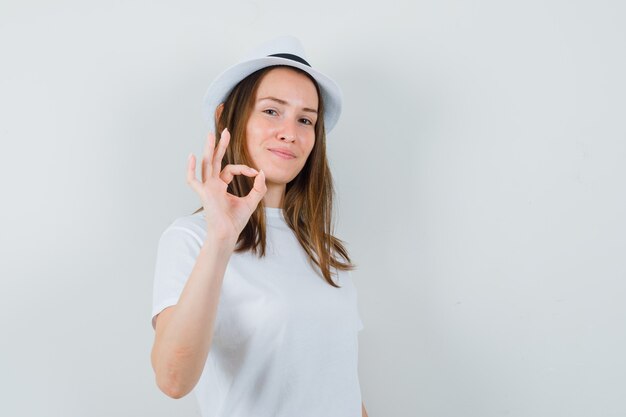 Young girl showing ok gesture in white t-shirt, hat and looking confident , front view.