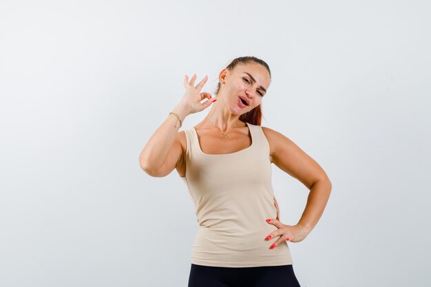 Young girl showing ok sign, holding hand on waist in beige top