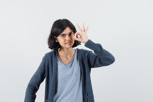 ライトグレーのTシャツとダークグレーのジップフロントフーディーでOKサインを見せて、かわいく見える少女。