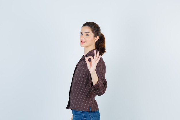 Free photo young girl showing ok sign, looking over shoulder in striped shirt, jeans and looking cheery , front view.