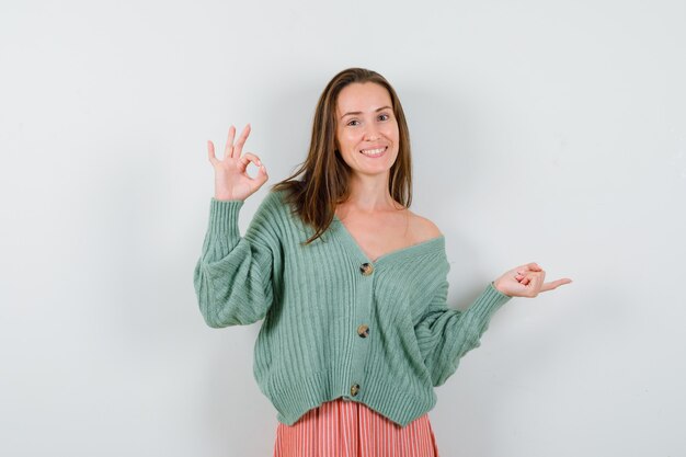 Young girl showing ok sign and pointing right with index finger in knitwear, skirt and looking cheery. front view.