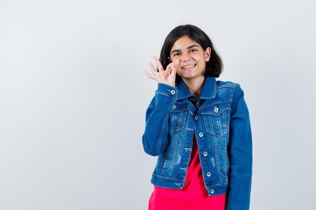 Young girl showing ok sign in red t-shirt and jean jacket and looking happy , front view.