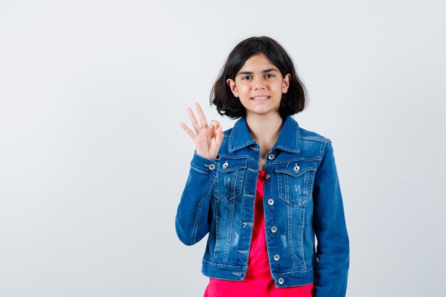 Young girl showing ok sign, smiling in red t-shirt and jean jacket and looking cheerful. front view.