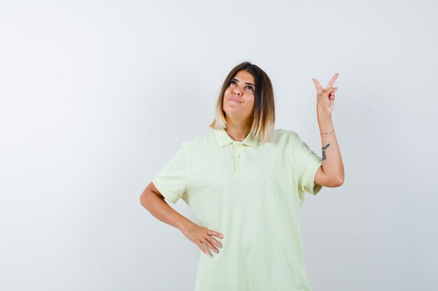 Young girl showing peace gesture, holding hand on waist in t-shirt and looking pensive , front view.