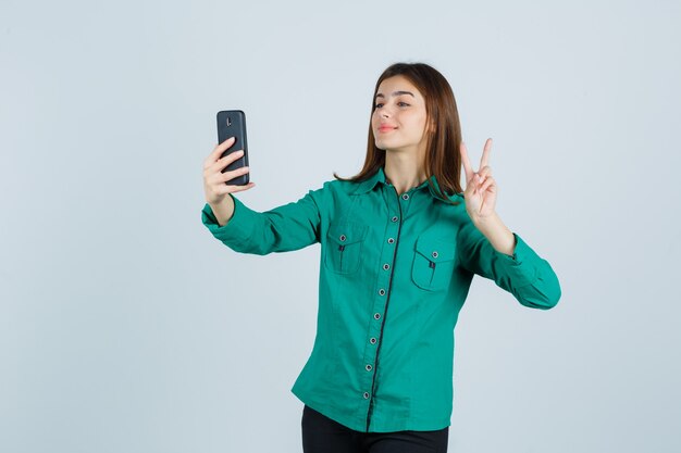 Молодая девушка показывает жест мира во время видеозвонка в зеленой блузке, черных штанах и выглядит мило, вид спереди.