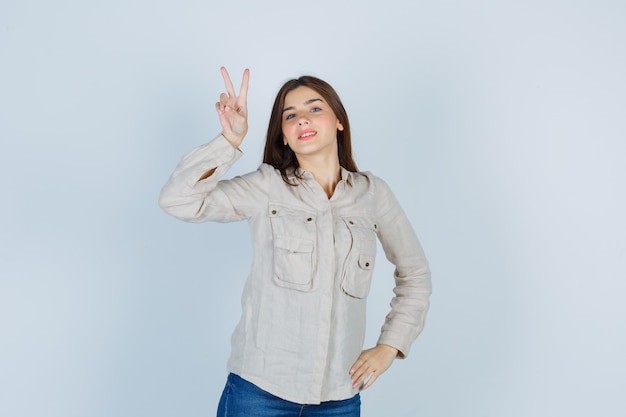 Молодая девушка показывает жест мира, с рукой на бедре в бежевой рубашке, джинсах и выглядит уверенно, вид спереди.