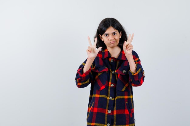 Young girl showing peace gestures in checked shirt and looking happy. front view.