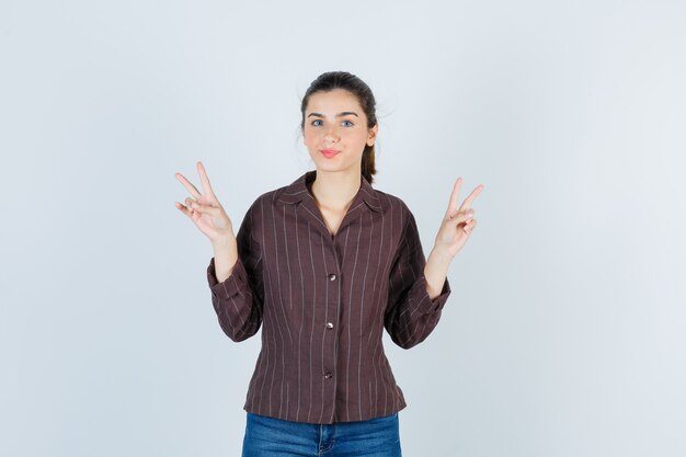 Молодая девушка показывает жесты мира в полосатой рубашке, джинсах и выглядит мило, вид спереди.