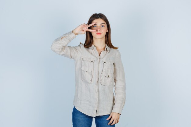 Молодая девушка показывает знак мира на глазу в бежевой рубашке, джинсах и выглядит серьезным, вид спереди.