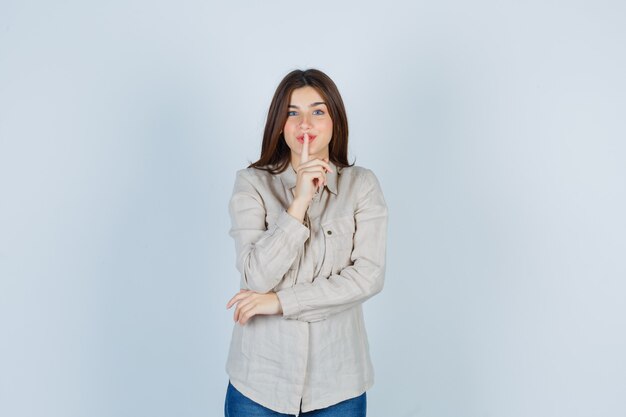 Young girl showing silence gesture in beige shirt, jeans and looking serious. front view.