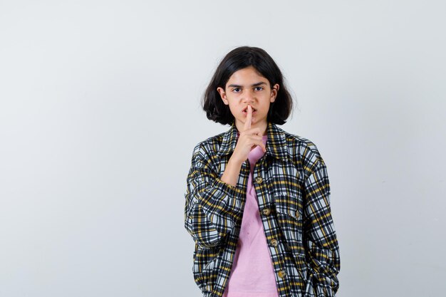 Young girl showing silence gesture in checked shirt and pink t-shirt and looking serious , front view.