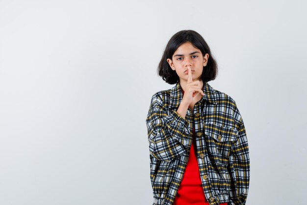 Young girl showing silence gesture in checked shirt and red t-shirt and looking cute , front view.