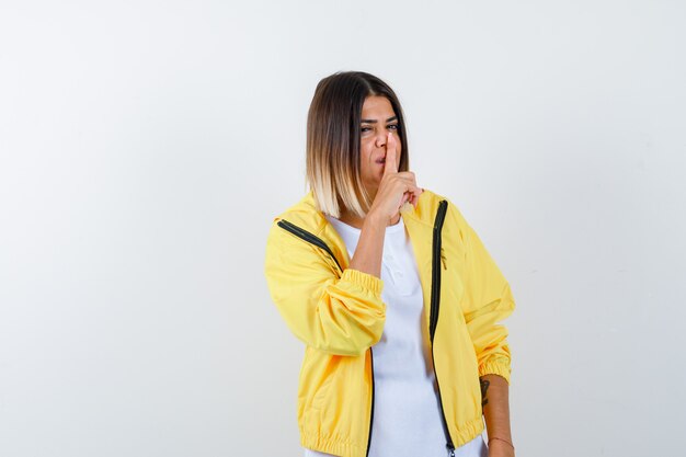 Young girl showing silence gesture in white t-shirt , yellow jacket and looking serious , front view.