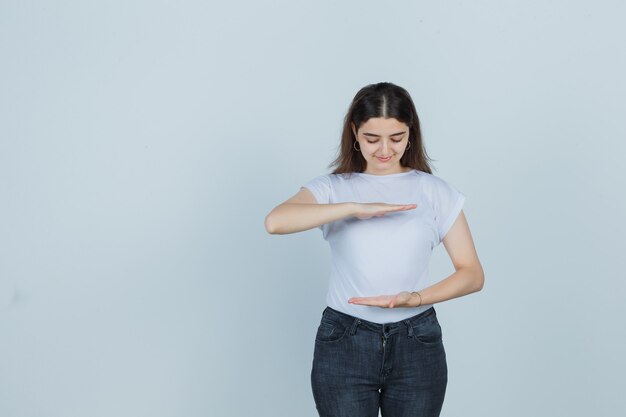 Молодая девушка показывает знак размера в футболке, джинсах и выглядит довольным, вид спереди.