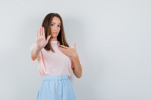 Young girl showing stop gesture while asking 'me?' in t-shirt, skirt and looking serious. front view.