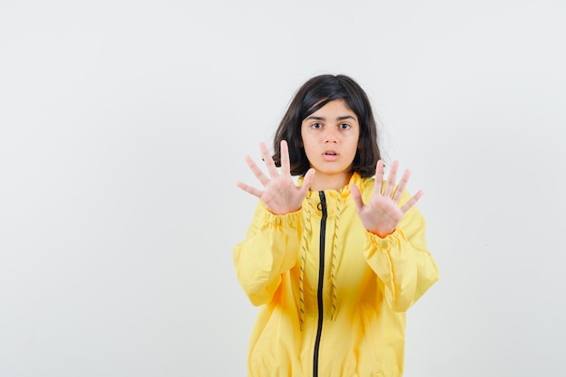 Young girl showing stop gesture with both hands in yellow bomber jacket and looking serious