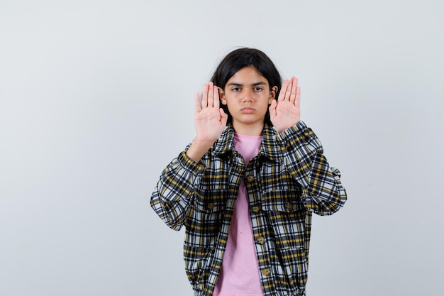 Young girl showing stop signs in checked shirt and pink t-shirt and looking serious. front view.