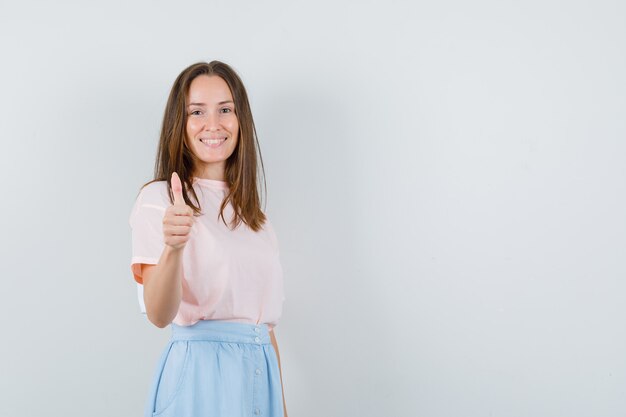 Young girl showing thumb up in t-shirt, skirt and looking pleased. front view.