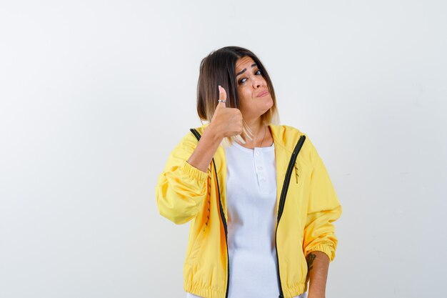 Молодая девушка показывает палец вверх в белой футболке, желтой куртке и выглядит впечатленным, вид спереди.