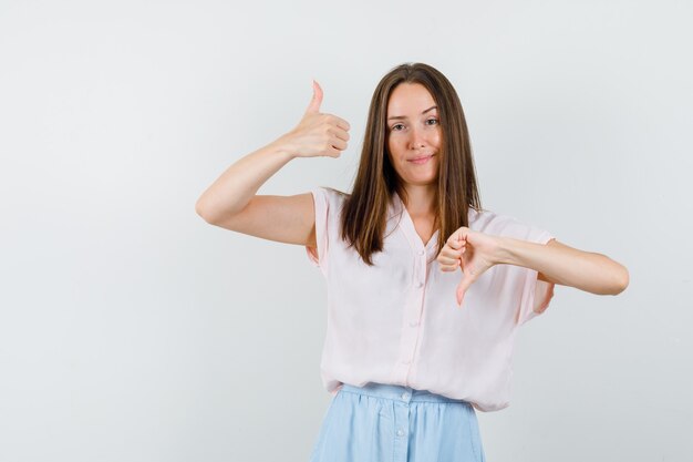 Tシャツ、スカートで親指を上下に見せて、賢明に見える、正面図の若い女の子。