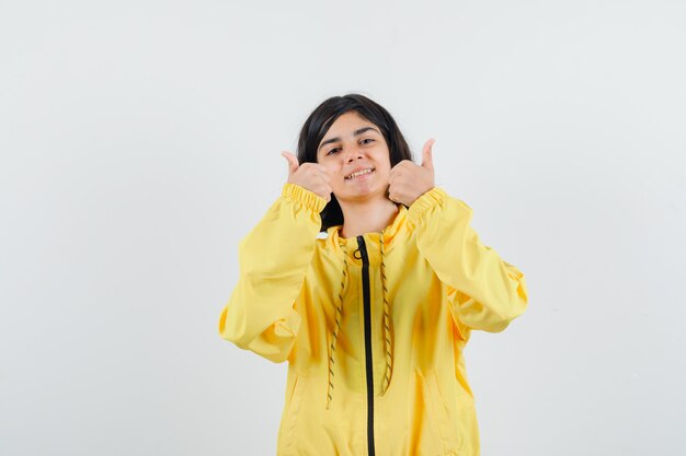 Young girl showing thumbs up with both hands in yellow bomber jacket and looking happy.