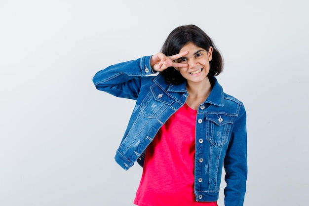 Young girl showing v sign on eye in red t-shirt and jean jacket and looking cute