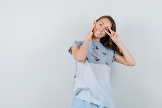 Young girl showing v-sign on eye in t-shirt, skirt and looking cheerful , front view.