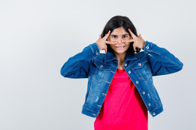 Young girl showing v signs on eye in red t-shirt and jean jacket and looking cute. front view.