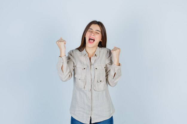 Молодая девушка показывает жест победителя в бежевой рубашке, джинсах и выглядит удачливым, вид спереди.