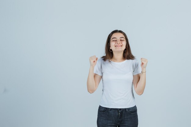 Молодая девушка показывает жест победителя в футболке, джинсах и выглядит удачливым, вид спереди.