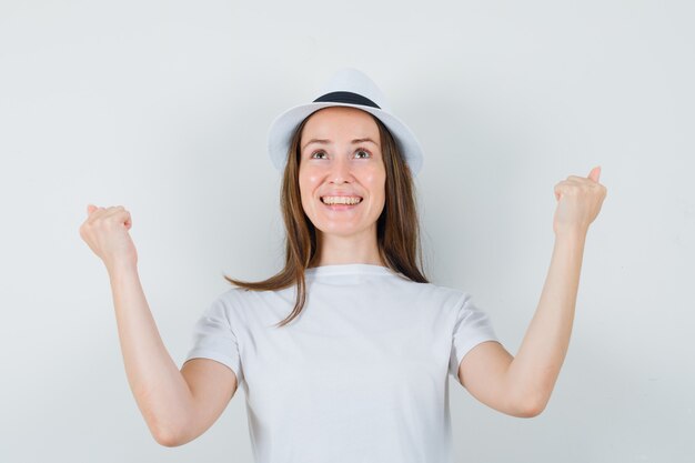 Молодая девушка показывает жест победителя в белой футболке и выглядит счастливой