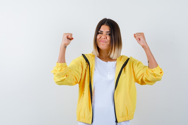 Young girl showing winner gesture in white t-shirt , yellow jacket and looking lucky , front view.